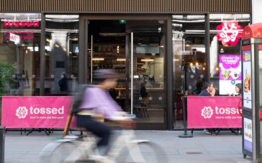 A person rides a bicycle past the entrance of a “tossed” restaurant, with pink signage and two outdoor tables visible on the pavement.