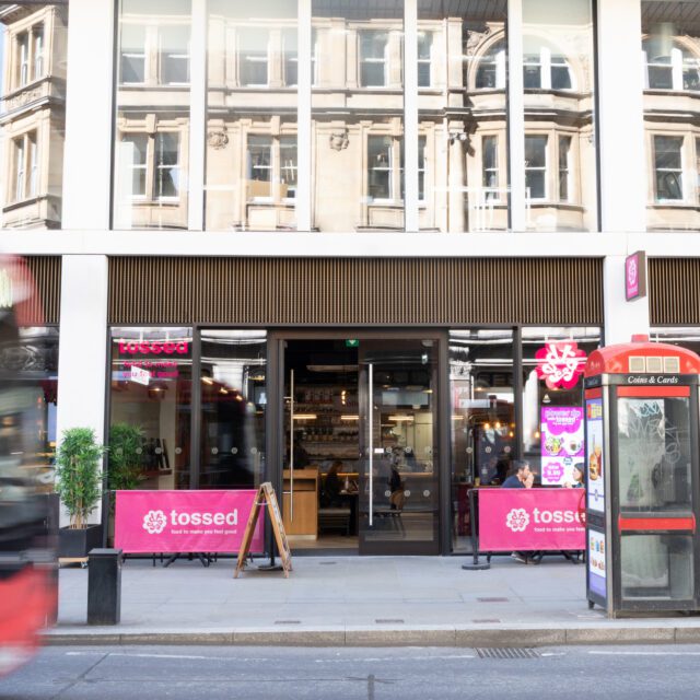 A red bus passes a restaurant called “tossed” with pink signage. A classic red phone box stands nearby, and pedestrians walk along the pavement in front of large city windows.