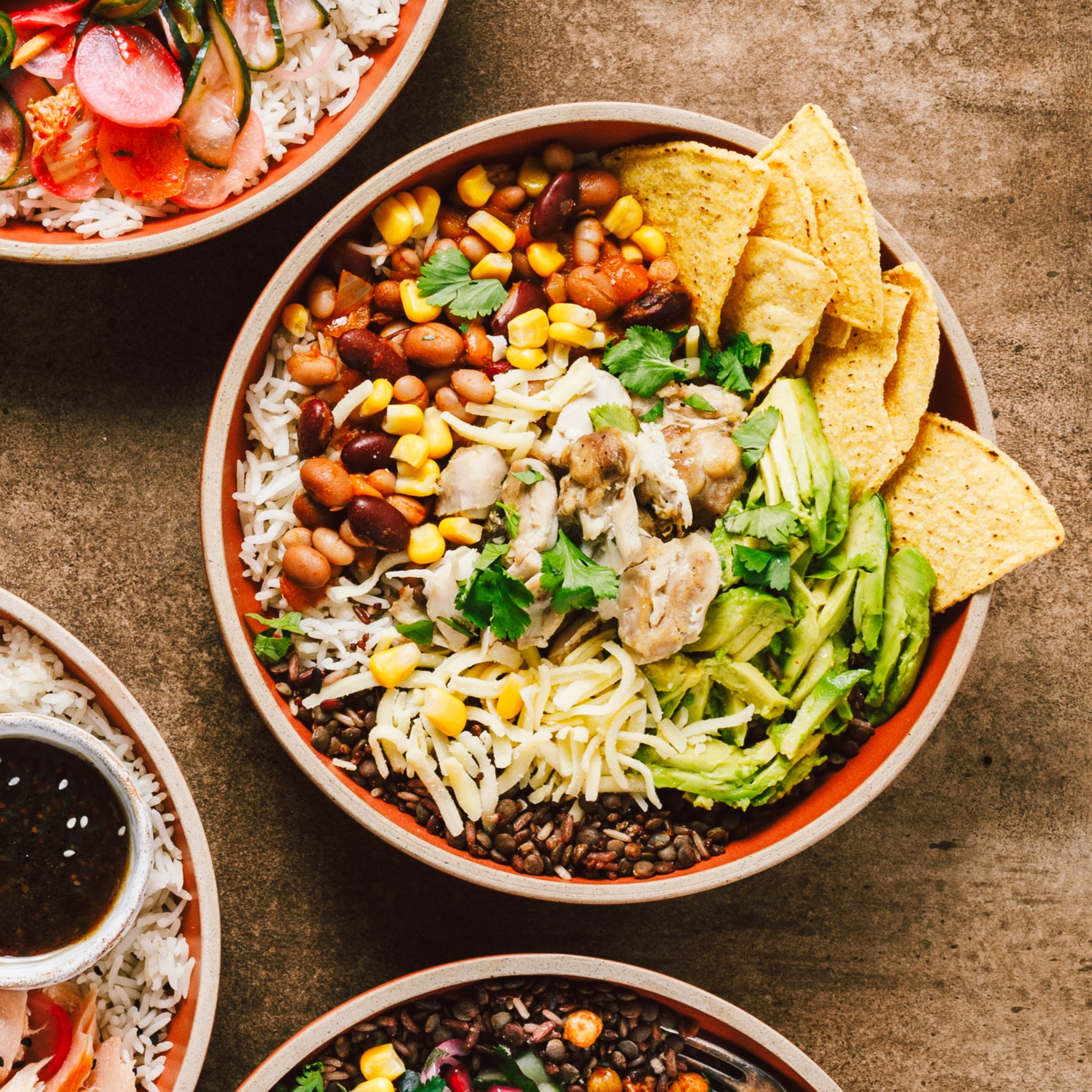 A colourful food burrito bowl with rice, beans, sweetcorn, grated cheese, sliced avocado, coriander, tortilla chips, and grilled chicken, surrounded by other bowls with various ingredients on a brown surface.