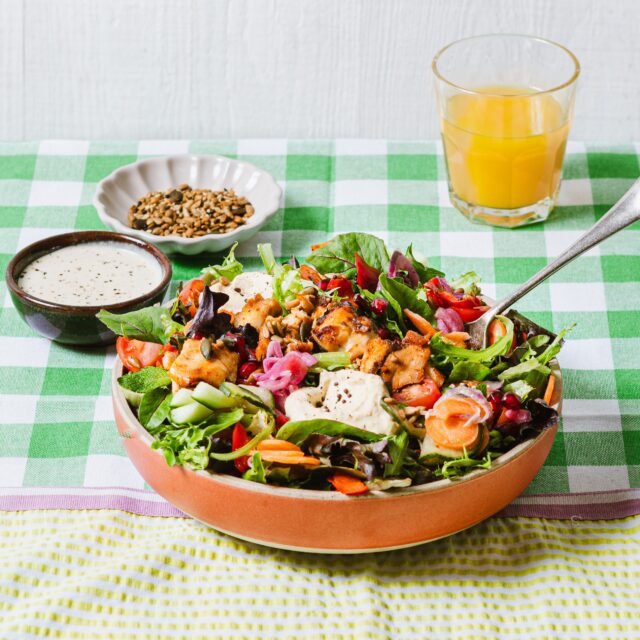 A colourful salad with grilled chicken, mixed greens, sliced carrots, cucumbers, pickled onions, and dressing in a bowl. Nearby are a dish of seeds, a bowl of dressing, and a glass of orange juice on a green checked tablecloth.