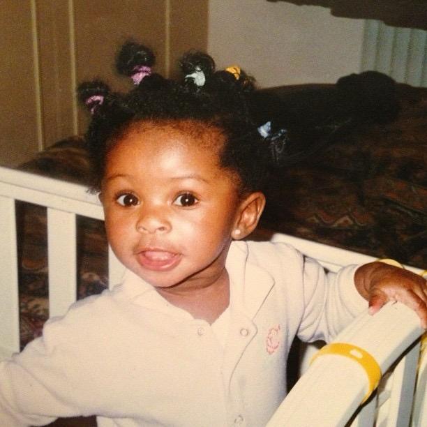 A toddler with multiple colourful hair bands stands in a cot, smiling and looking at the camera. She wears a light-coloured babygrow and holds onto the cot rail.