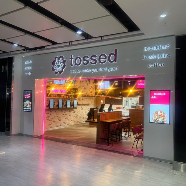 The shopfront of a Tossed restaurant, featuring a sign listing food options like salads, wraps, and bowls, with a view into an open interior with bar seating.